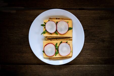 Sandwich made from rye bread, hard cheese, radish, arugula, served on a white plate against a dark wooden background. Symmetry. Copy space. Flat lay, top view. Close-upの写真素材