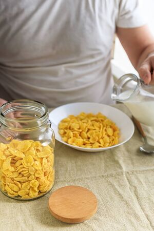 Cropped man pours milk into cornflakes in a white plate on a plain rough tablecloth. View from above. Selective focus. Concept, simple fast american healthy breakfastの写真素材