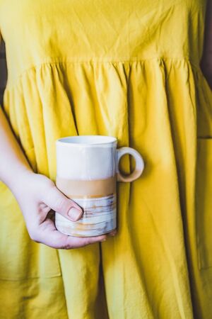 Cropped young white woman in a simple yellow linen dress holds in her hands a clay ceramic cup of an original handmade shape. Copy space. Trend.の写真素材
