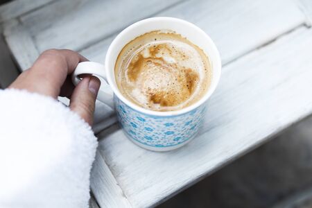 cropped female hand and cappuccino coffee in a cup on a white wooden table. Top view, copy space.の写真素材