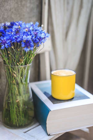 on the balcony there is a white wooden table with a thick book, a yellow cup of coffee and a vase with cornflowersの写真素材