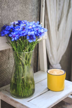 on the balcony is a white wooden table with a yellow cup of coffee and a vase with cornflowers.の写真素材