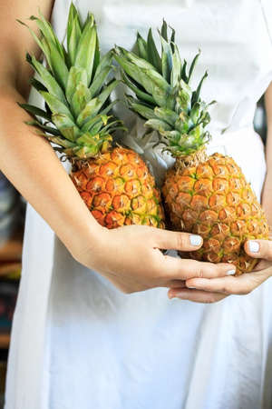 cropped young woman in a white cook apron holds in her hands two ripe and juicy pineapples.の写真素材