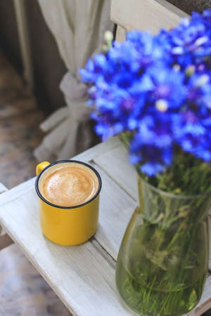 on the balcony is a white wooden table with a yellow cup of coffee and a vase with cornflowers.の写真素材