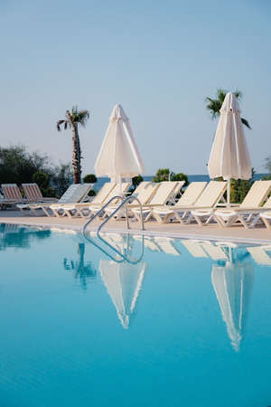 View of the pool, sun loungers and parasols among lush green palms and bushes in the soft morning...