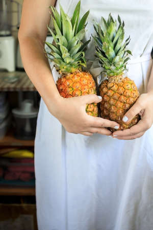 cropped young woman in a white cook apron holds in her hands two ripe and juicy pineapples.の写真素材