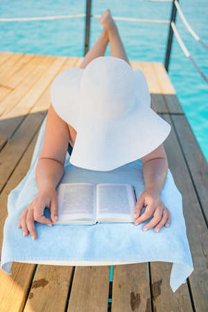 Beautiful young woman reading book while relaxing on sun lounger near swimming poolの写真素材