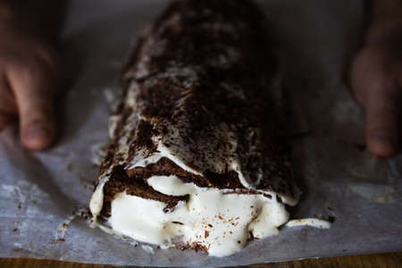 A traditional Christmas chocolate roll-log with cream, against the background of Christmas tree lights on a dark background, is held by a manの写真素材
