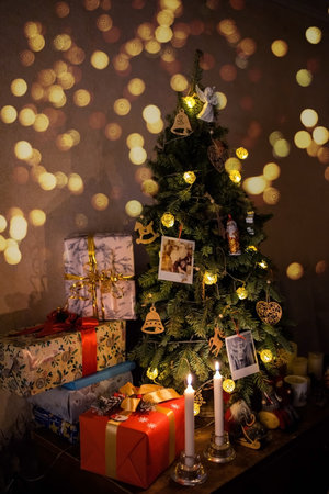 beautiful wrapped boxes with gifts near the Christmas tree, there are candles, lights nearby, family photos and a garland are hanging on the treeの写真素材