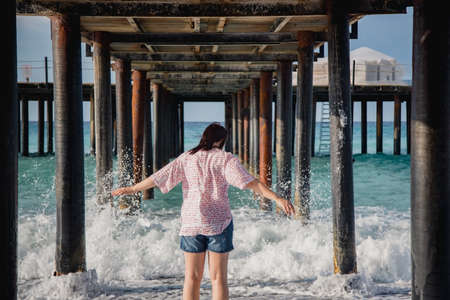 Turkey, Mediterranean Sea. Young woman turned away from the camera, running away from the waves under a wooden pier.の写真素材