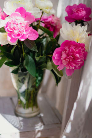 a bouquet of fresh white and pink peonies in a transparent glass vase with beautiful shadows near a white curtain tulle, in blurの写真素材