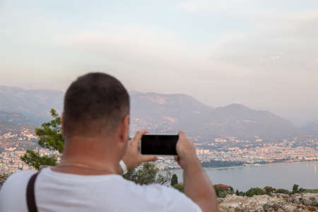 Unrecognizable young dark-haired man origin, from behind taking a horizontal photograph of the beach with his smartphone. Sunny and well-lit summer day, light and blue colorsの写真素材