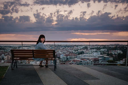 A young Caucasian woman sits on a bench and looks at the panorama of the city of Manavgat, Turkey at sunset.の写真素材