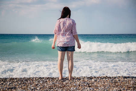 young woman in shorts and a shirt stands on a pebble beach and looks at the waves of the mediterranean sea, rear view, hair fluttering in the windの写真素材
