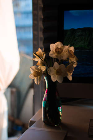 on a desktop with a monitor and keyboard, in a home office, there is a stained glass vase with fresh flowers of yellow daffodils. Concept, enjoyable work online remotely.の写真素材