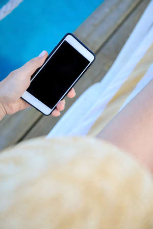 female hand holding a phone - smartphone on the background of a tanned leg on a blue sea lounger.の写真素材