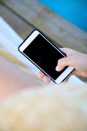 female hand holding a phone - smartphone on the background of a tanned leg on a blue sea lounger.の写真素材