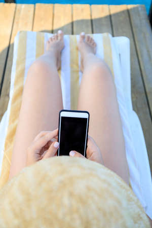 female hand holding a phone - smartphone on the background of a tanned leg on a blue sea lounger.の写真素材