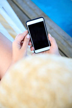 female hand holding a phone - smartphone on the background of a tanned leg on a blue sea lounger.の写真素材