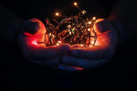 male hands holding a Christmas garland with lights on a dark background with copy space for text. Retro effect, close-up. Winter, postcard templateの写真素材