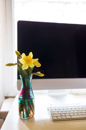 on a desktop with a monitor and keyboard, in a home office, there is a stained glass vase with fresh flowers of yellow daffodils. Concept, enjoyable work online remotely.の写真素材