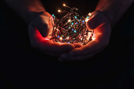 male hands holding a Christmas garland with lights on a dark background with copy space for text. Retro effect, close-up. Winter, postcard template.の写真素材