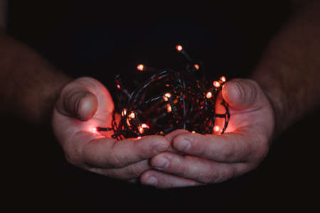 male hands holding a Christmas garland with lights on a dark background with copy space for text. Retro effect, close-up. Winter, postcard template.の写真素材