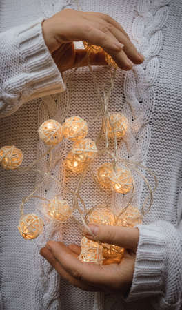 lights in the palms. Female hands hold a garland with wicker round balls. Girl in a white sweater with Christmas lights in her hands, warm New Year mood, soft focusの写真素材