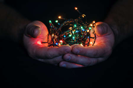 male hands holding a Christmas garland with lights on a dark background with copy space for text. Retro effect, close-up. Winter, postcard template.の写真素材