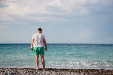 attractive young adult man stands with his back on the shore of the mediterranean sea with waves. copyspace.の写真素材