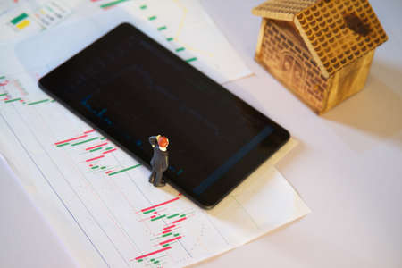 miniature business man with stock exchange graph data on white paper and blurred tablet with blurred...
