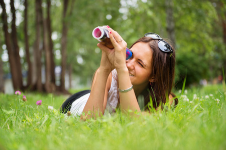Young brunette woman enjoying the lying on the green grass in the garden and looking through rolled-up magazineの写真素材