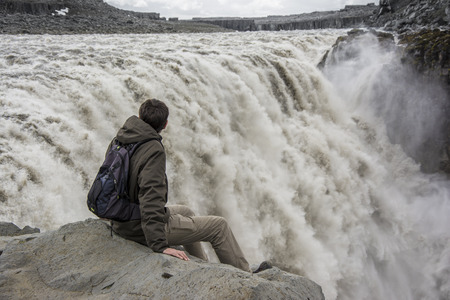 Young man on the edge contemplating Detifoss waterfall. This waterfall is one of the best attraction and the most powerful waterfall on Iceland and in the whole Europe.の写真素材