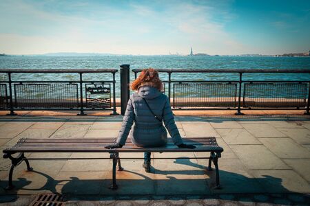 Curly brunette woman sitting on a bench and looking at the Statue of Libertyの写真素材