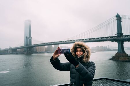 Tourist woman with warm clothes taking a selfie with the Manhattan Bridge on the backgroundの写真素材