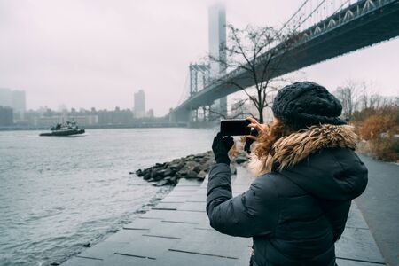 Tourist woman with warm clothes taking a picture of Manhattan Bridgeの写真素材