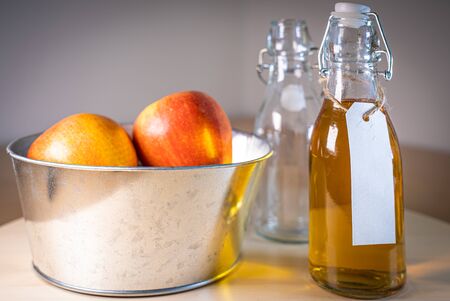 Close up view of glass bottle with a blank tag of apple juice or cider and apples in basket on wooden tabletopの写真素材