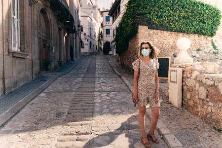 Beautiful curly brunette tourist with a surgical mask during Covid-19 in the Mediterranean village of Sitges, Barcelona provinceの写真素材