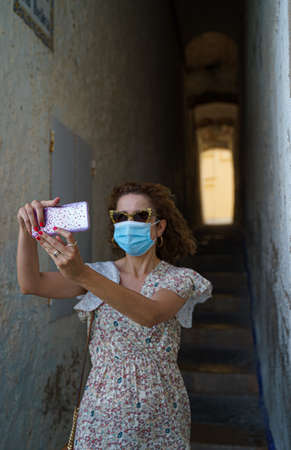 Beautiful curly brunette tourist with a surgical mask taking a sitges in the correlo de la rectoria in Sitges, spain.の写真素材