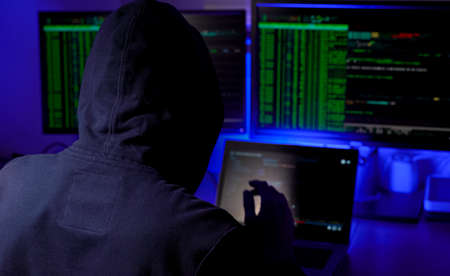 man in a hood working as a hacker at the computer in the dark room at night, hacking the system and laundring secret information.の写真素材