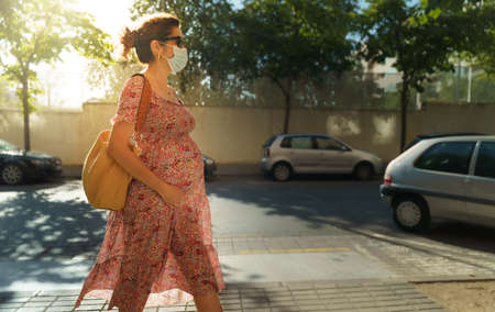 Pregnant curly brunette wearing a face mask and walking in a city street.の写真素材