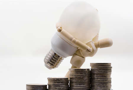 wooden doll holding a light bulb on his shoulders with a rising coin graph in the foreground . Concept of rising electricity pricesの写真素材