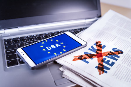 Smartphone with DSA and EU flag on screen over a newspaper with Fake news and a laptop. Fake news, HOAX conceptの写真素材