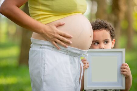 The little boy is happy with the pregnant mother. The garden is full of green trees.