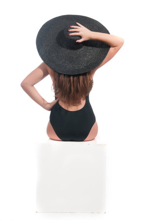 Young woman in black swimsuit and black hat sitting on white cubeの写真素材