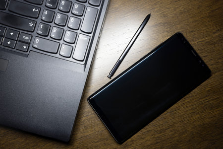 A black smartphone with a black laptop and a black pen are on the brown wooden working table.の写真素材
