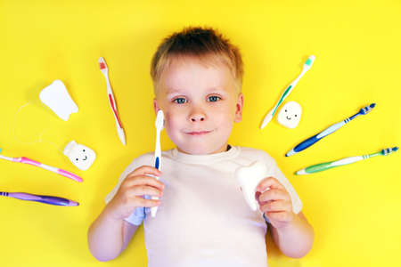Cute boy holding a toothbrush and a tooth toy surrounded by toothbrushes on yellow background top...