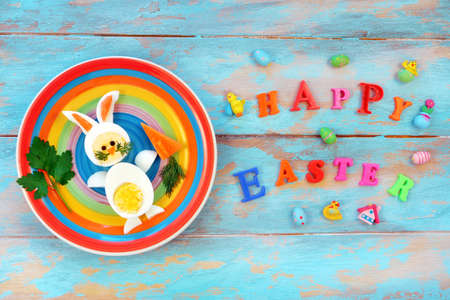 Easter breakfast idea for kids. Bunny rabbit made of boiled eggs on multicolored plate, blue wooden background, inscription Happy Easter, top viewの写真素材