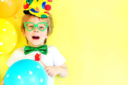 Funny laughing child clown on yellow background with multicolored balloons, copy space. April fools day celebration concept. Birthday party concept.の写真素材