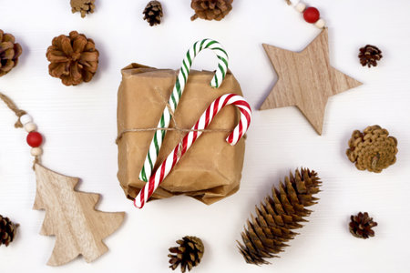 Christmas gift box in craft paper surrounded by wooden christmas decorations and fir cones , white...